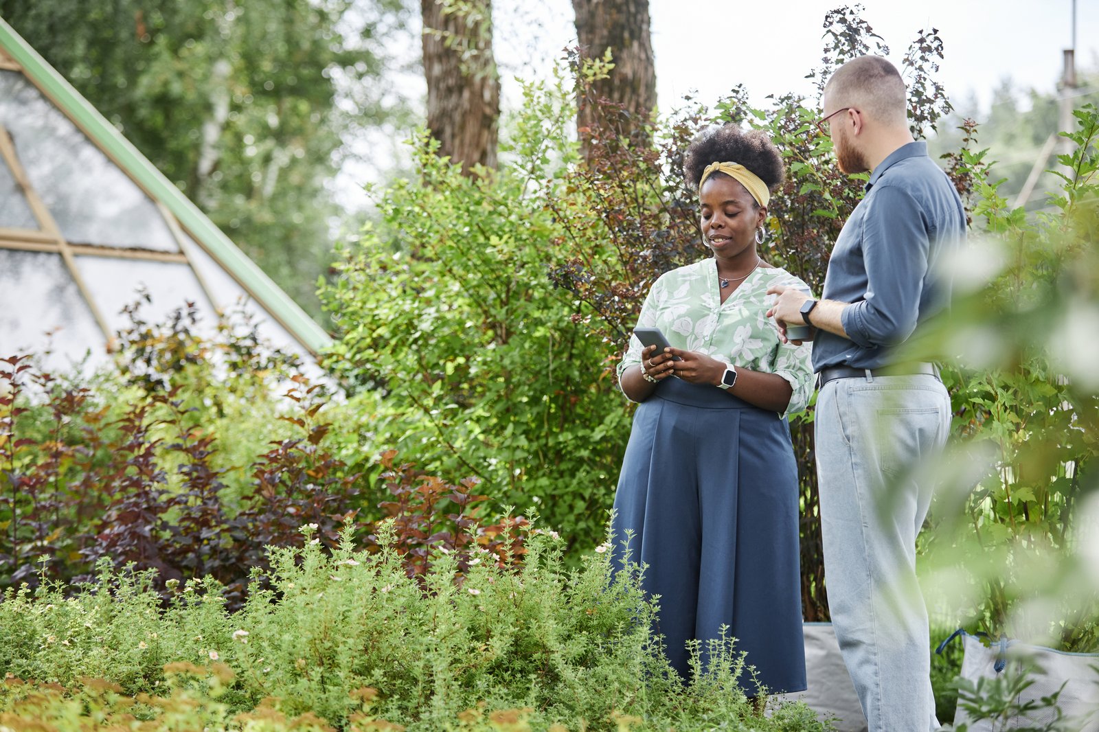 Positive African American female landscape designer using mobile phone while discussing with male client garden project providing man with professional assistance captured against lively greenery