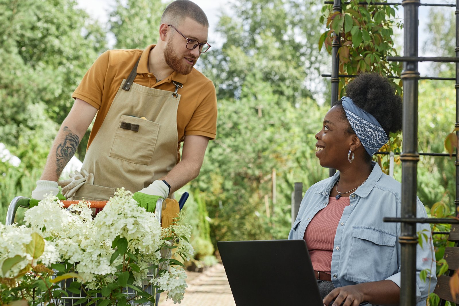 Medium shot of cheerful African American female landscape designer enjoying working on laptop outside in green garden, while chatting with male plant nursery worker passing by with flowers on cart