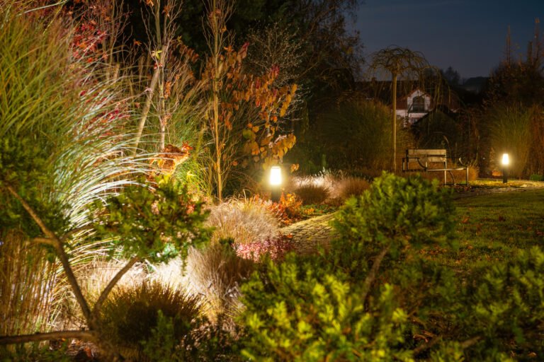 A garden path is illuminated by soft lights, surrounded by autumn foliage and a cozy bench under the evening sky, creating a serene atmosphere.